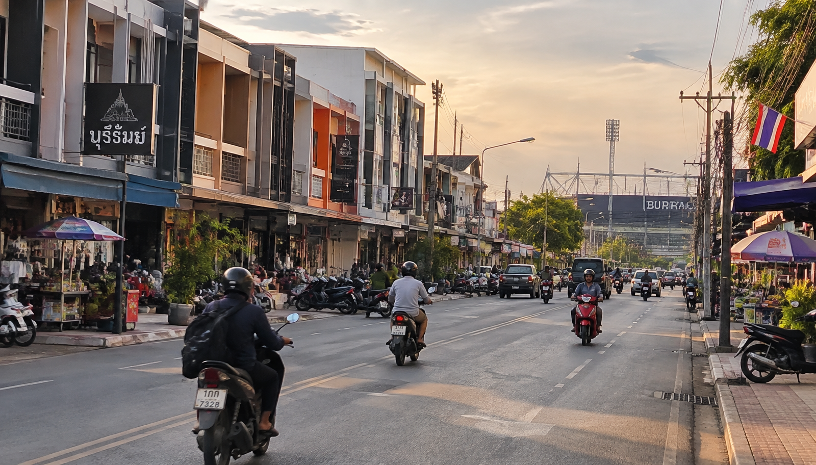 ถนนในเมืองบุรีรัมย์ยามเย็น มีตึกแถว รถจักรยานยนต์ และสนามกีฬาเห็นอยู่ไกล ๆ